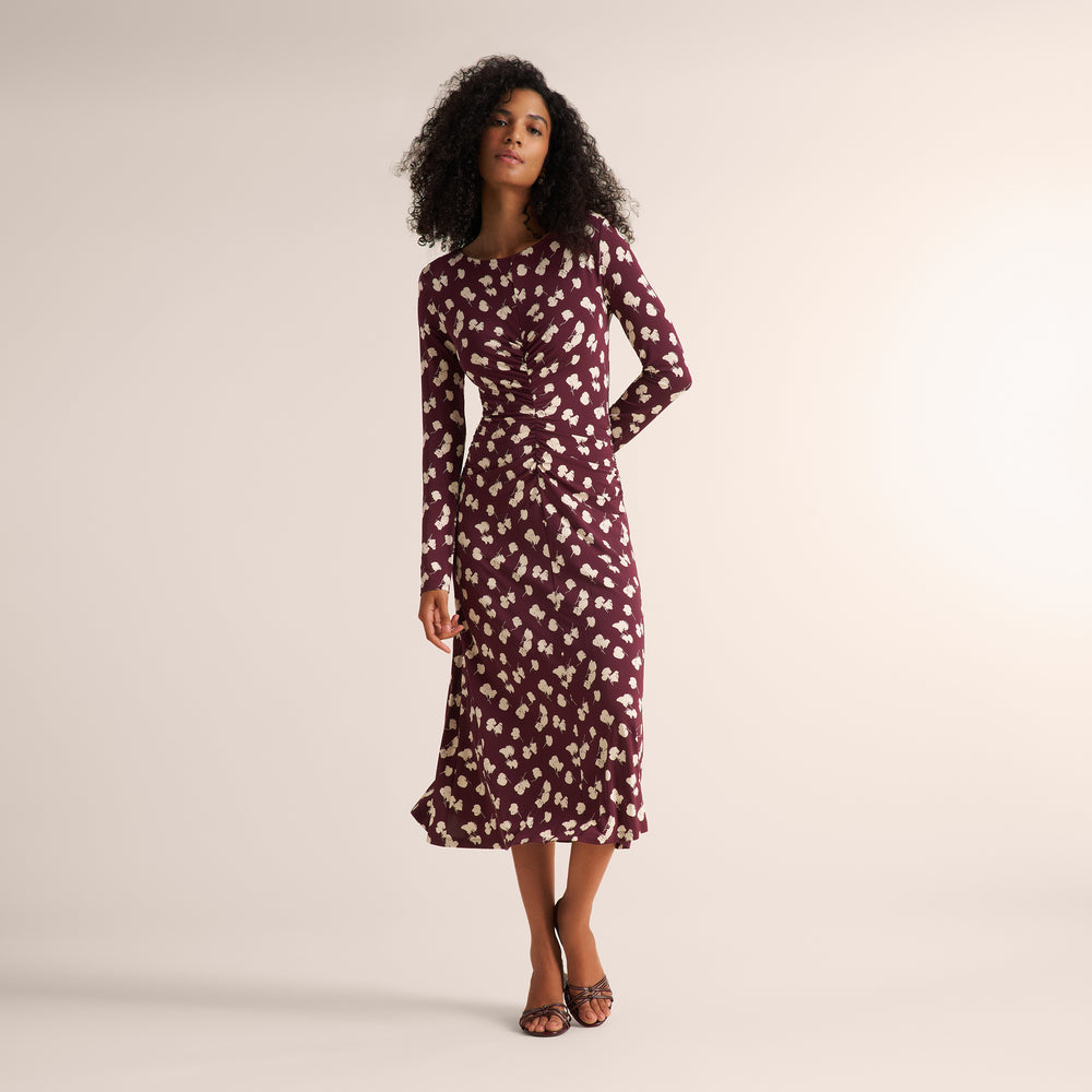 A woman modeling a long-sleeve midi dress with a burgundy and cream floral pattern, standing against a plain beige studio background.