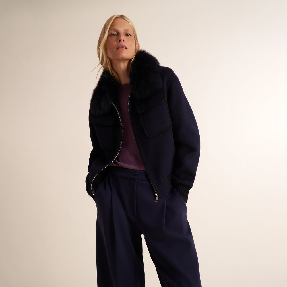 A woman modeling a dark navy blue jacket with a black fur collar, paired with matching navy trousers and a plum purple top, against a plain beige studio background.