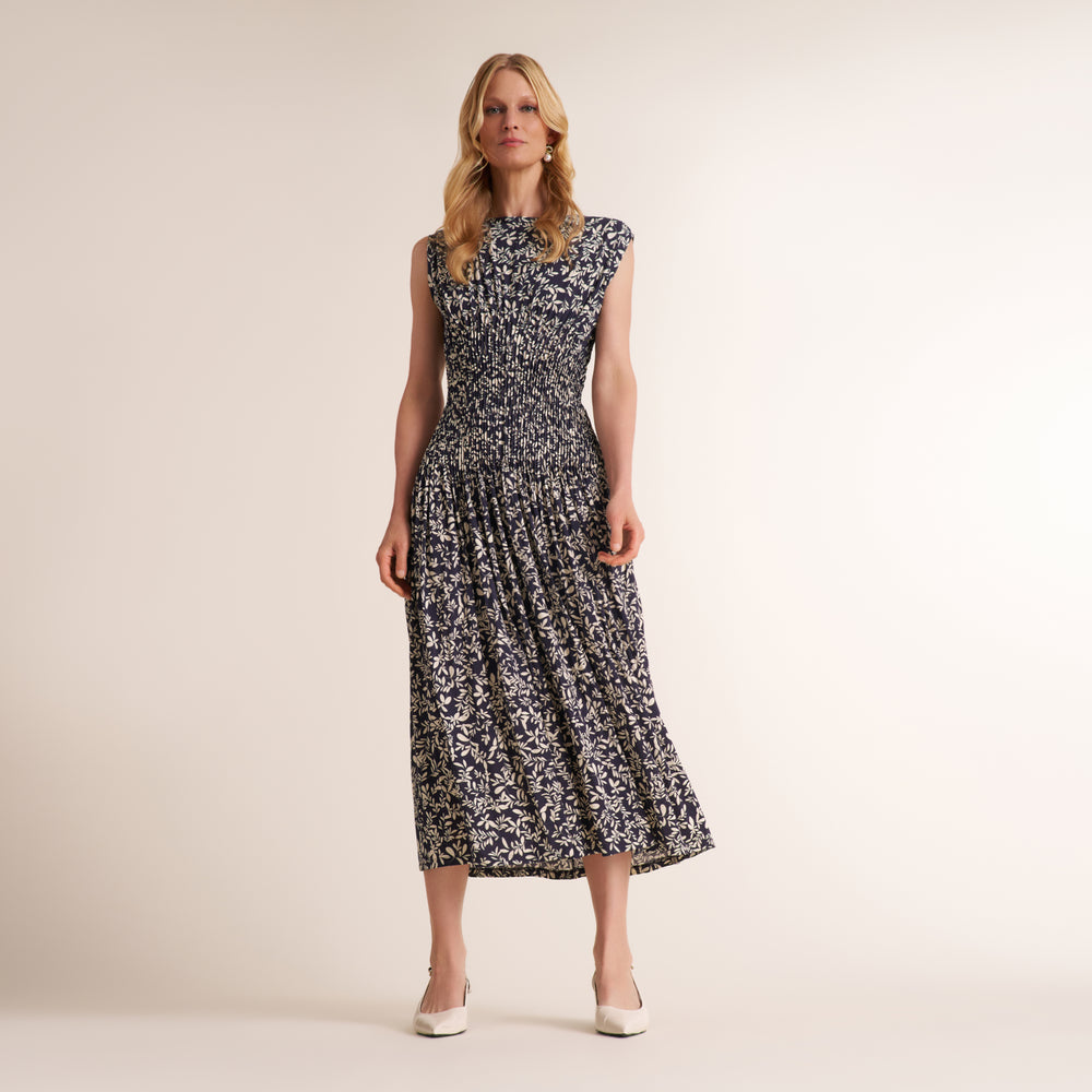A woman modeling a sleeveless midi dress with a black and white abstract floral pattern, standing against a plain beige studio background.