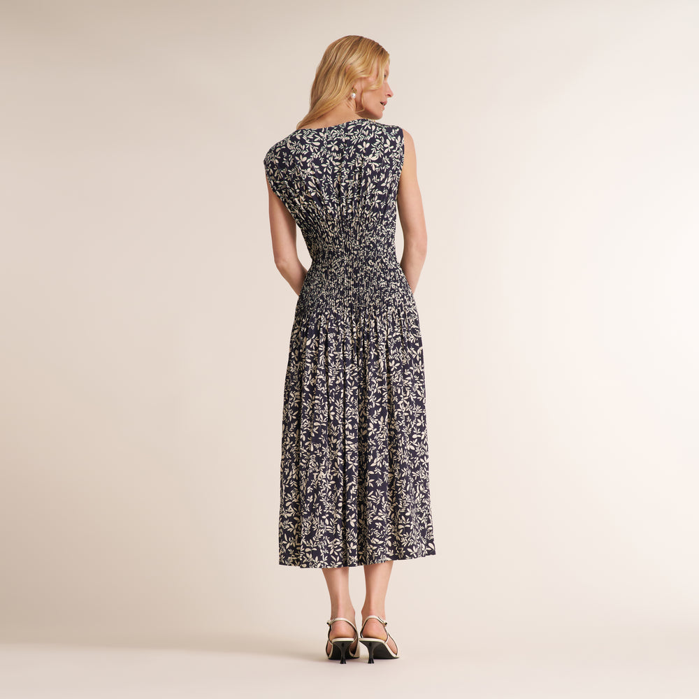 A woman stands with her back to the camera wearing a sleeveless midi dress with a black and white floral pattern, paired with black ankle-strap heels, against a solid beige studio background.