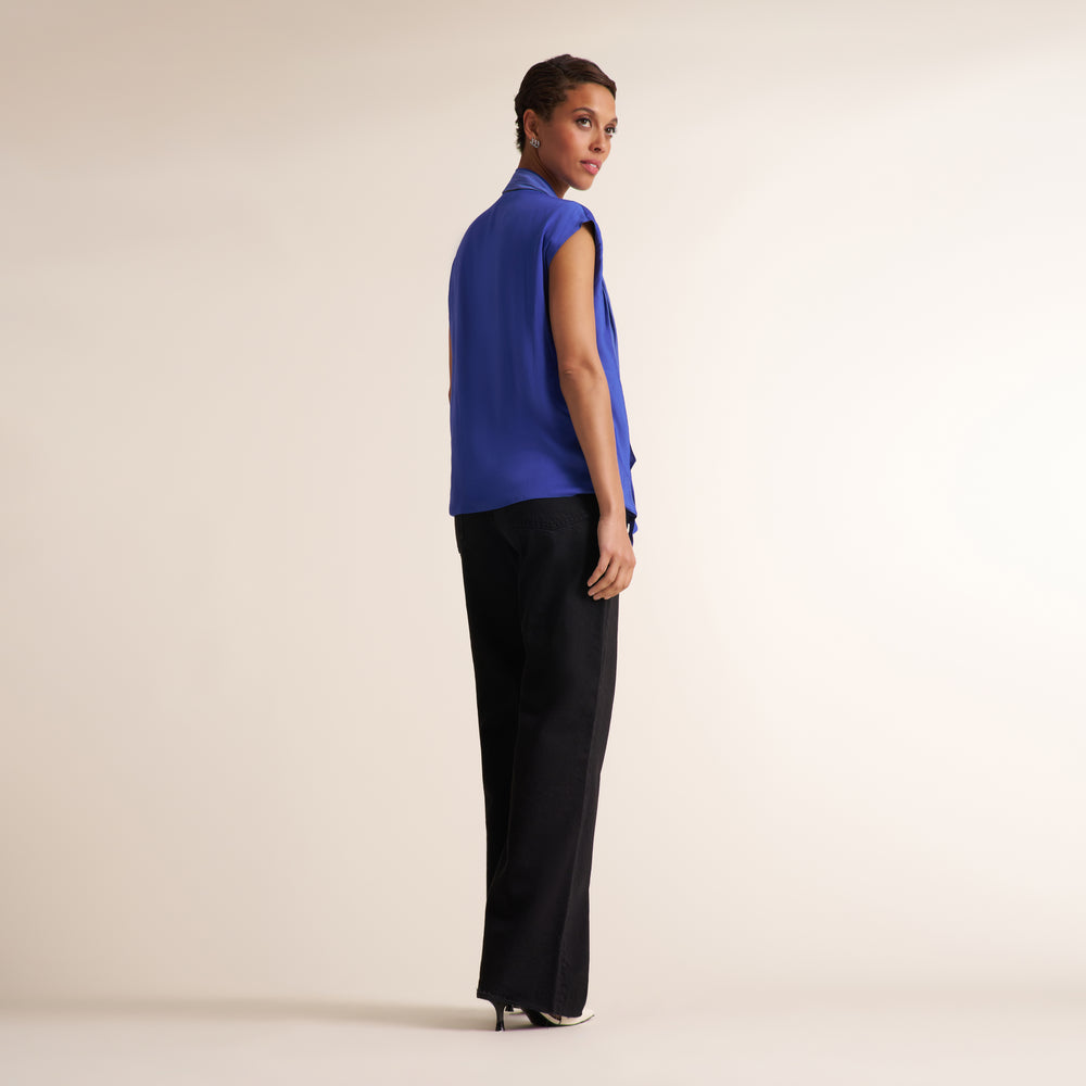 A model is shown from a side-back angle wearing a sleeveless royal blue vest and black wide-leg trousers against a plain beige studio background.
