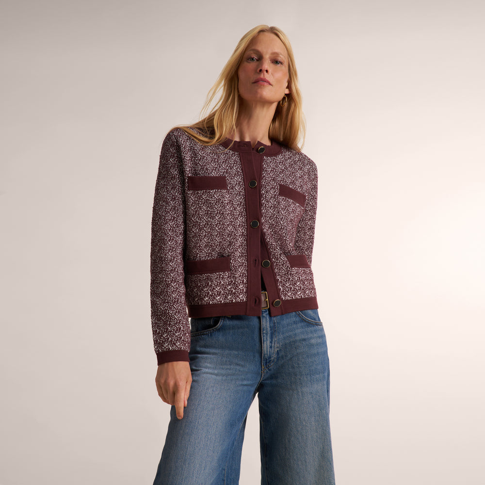 A woman modeling a cropped tweed jacket with a textured burgundy and cream pattern, paired with blue jeans, against a neutral studio background.