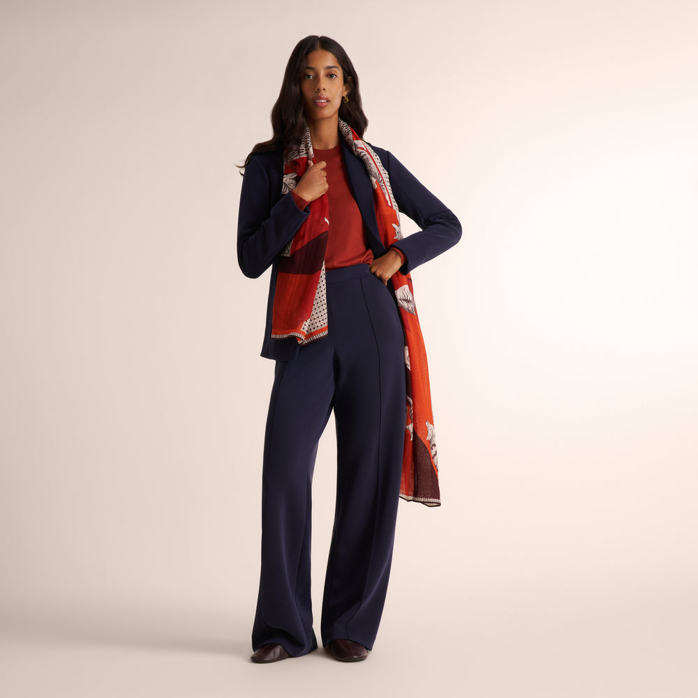 A woman models a navy blue two-piece suit with a red top and a red, orange, and white patterned scarf, standing against a solid light beige studio background.