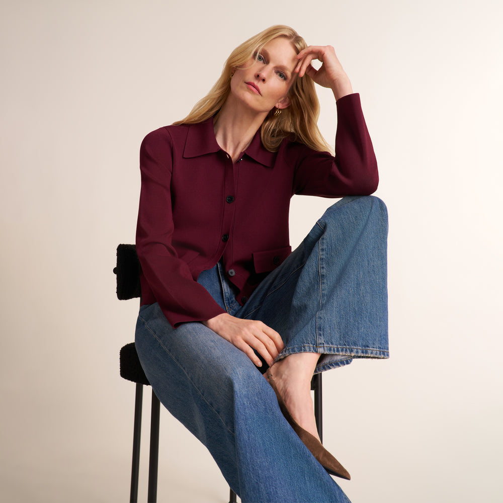 A woman sits on a black stool wearing a long-sleeve burgundy button-up shirt and wide-leg blue jeans against a plain beige studio background.