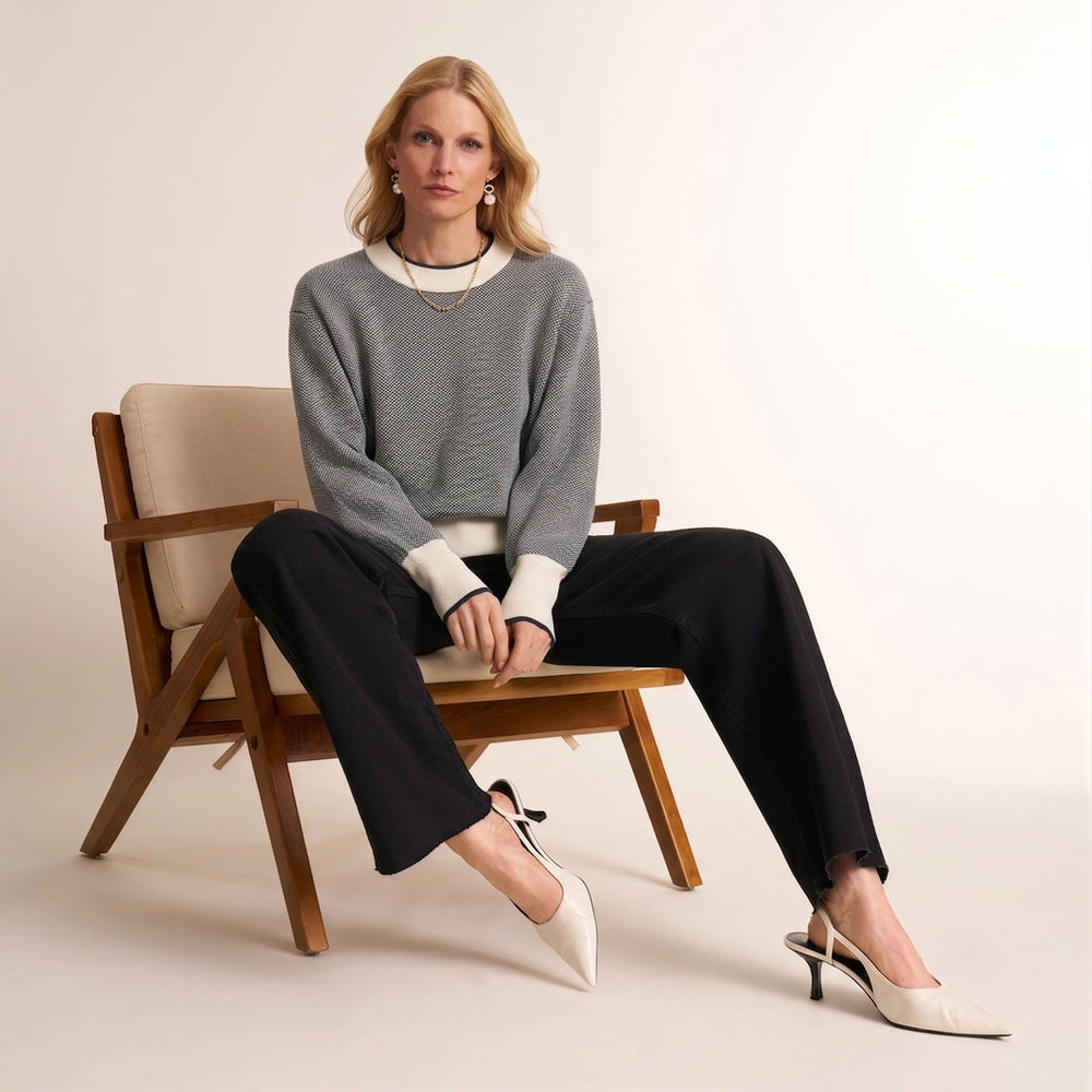 A woman sits on a wooden chair wearing a gray knit sweater with navy blue trim, black wide-leg pants, and white slingback heels against a plain light beige studio background.