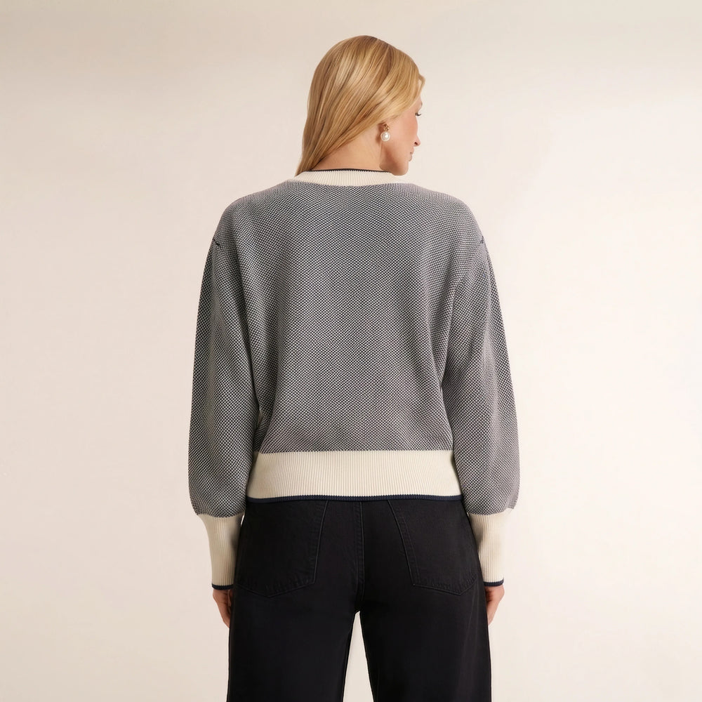 A woman shown from the back wearing a gray textured knit sweater with cream ribbed trim at the collar, hem, and cuffs, paired with dark pants, against a plain off-white studio background.