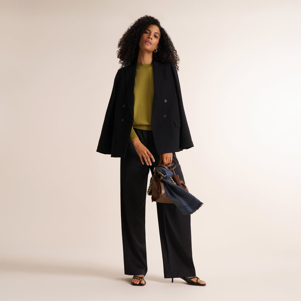 A model stands against a warm beige studio background wearing a black double-breasted blazer, black wide-leg trousers, and an olive green top. She is holding a brown leather handbag with a blue patterned scarf.