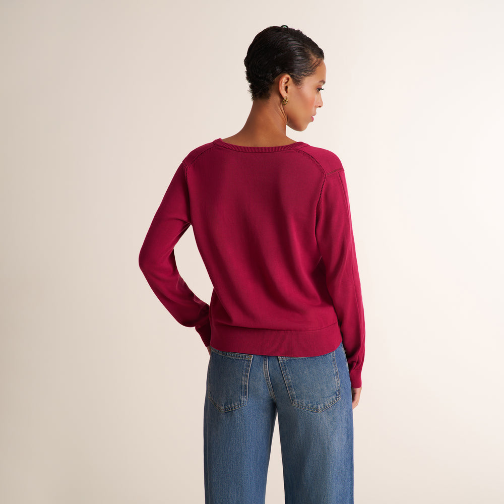A woman shown from the back wearing a long-sleeve crewneck sweater in deep burgundy and blue jeans, standing against a plain light beige studio background.