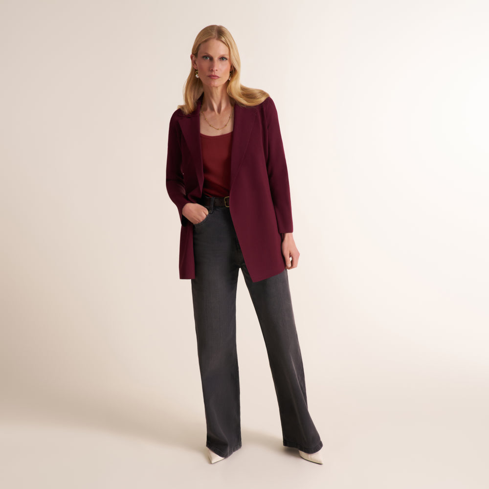 A woman models a burgundy open-front blazer over a matching burgundy top, paired with dark charcoal gray wide-leg trousers and white pointed-toe heels, against a solid off-white studio background.