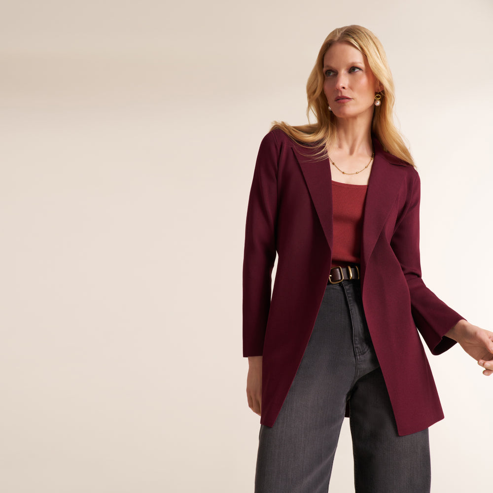 A woman modeling a long-sleeve open-front blazer in deep burgundy, worn over a matching burgundy top with dark gray jeans, against a plain light beige studio background.