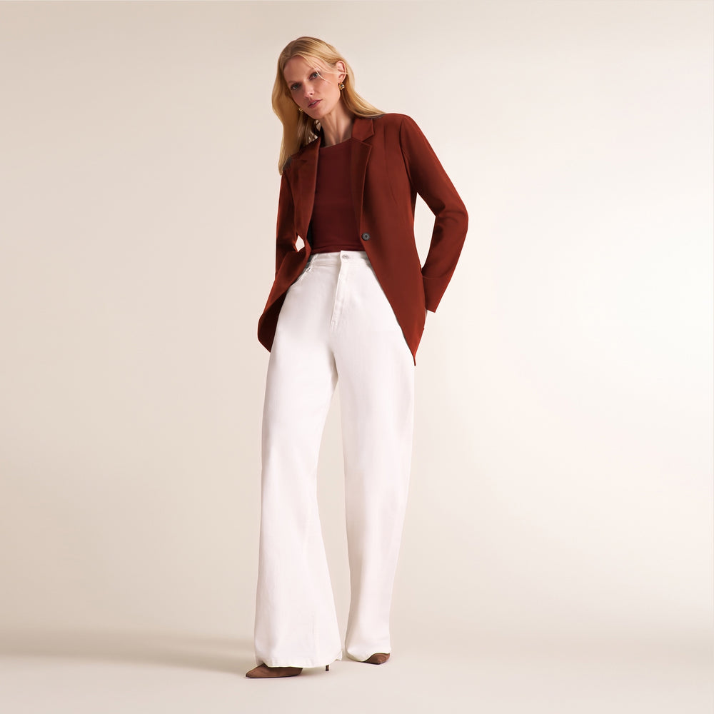 A woman modeling a rust-brown blazer over a matching rust-brown top and high-waisted white wide-leg trousers, standing against a solid light beige studio background.