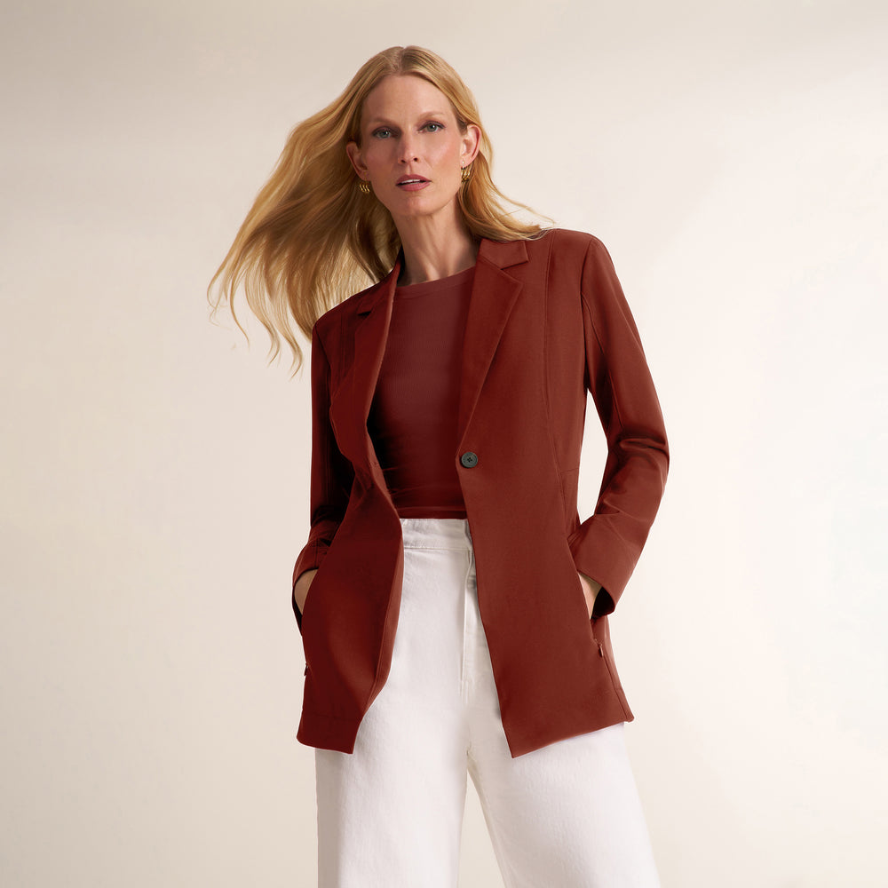 A woman modeling a rust-brown tailored blazer over a matching rust top with white pants, photographed against a plain warm off-white studio background.