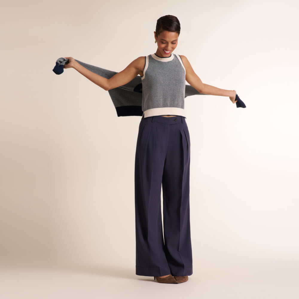 A woman modeling a sleeveless gray knit top with a white crew neck and a navy wide-leg pant, holding a navy cardigan open with both arms against a solid light beige background.