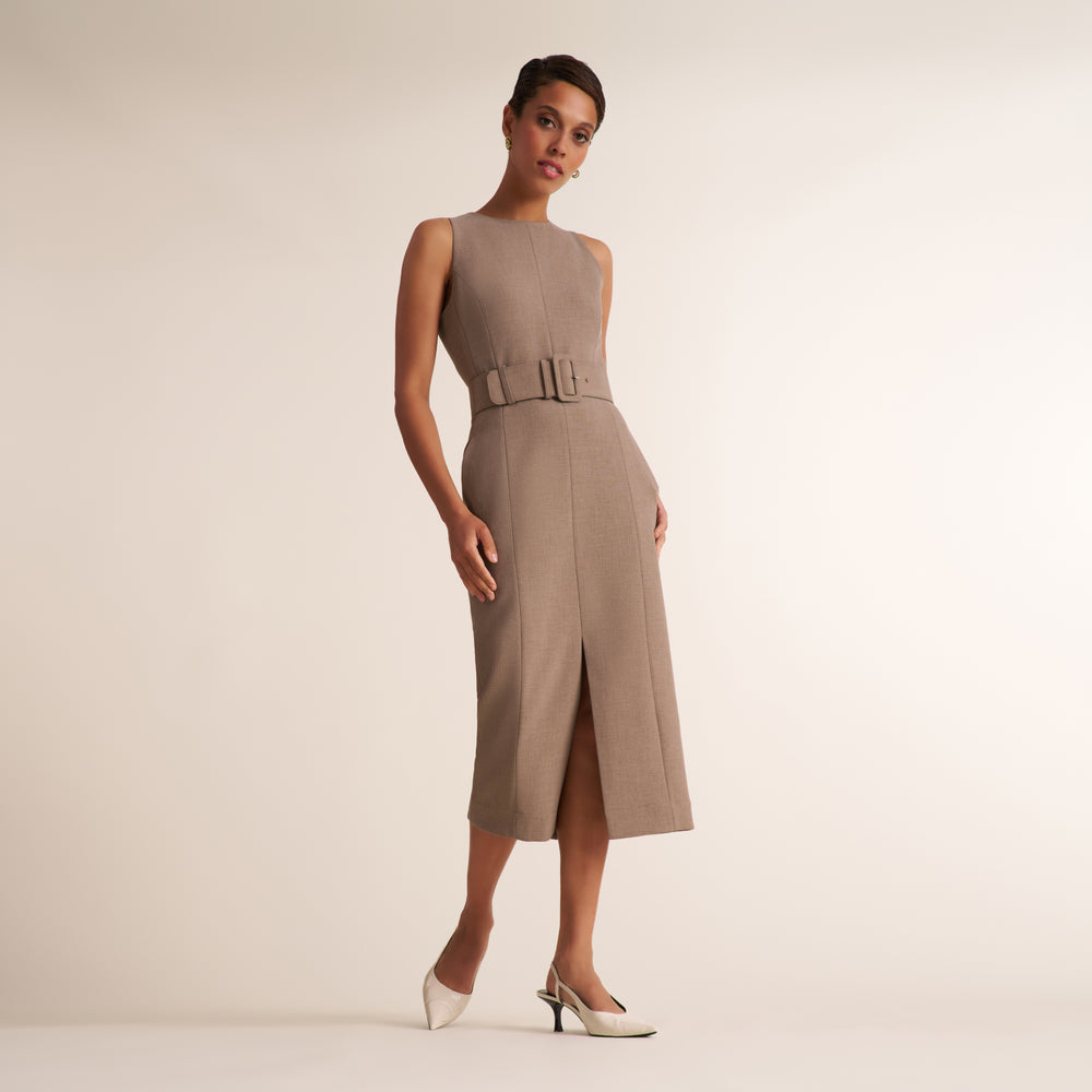 A model wears a sleeveless taupe midi dress with a matching belt and double buckle detail at the waist, paired with white pointed-toe heels, against a solid light beige studio background.