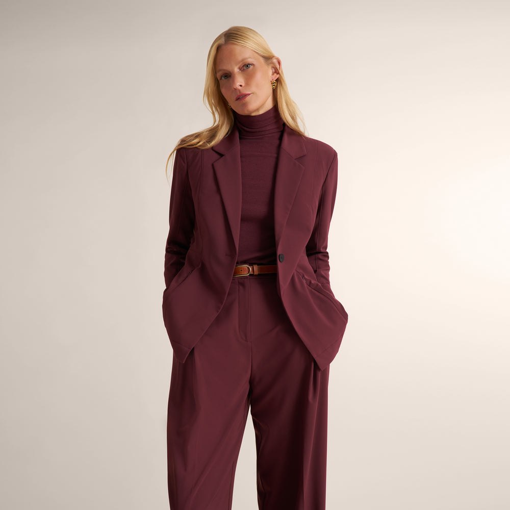 A woman modeling a tailored burgundy blazer and matching trousers with a matching turtleneck, cinched at the waist with a brown belt, against a plain light beige studio background.