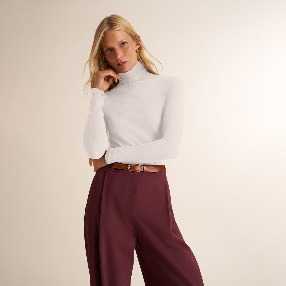 A blonde model wearing a white long-sleeve mock neck top and high-waisted burgundy wide-leg trousers with a brown leather belt, posing against a warm beige studio background.