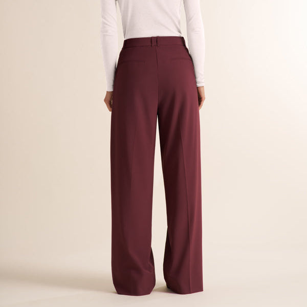 A woman shown from the back wearing a white long-sleeve mock neck top and high-waisted, wide-leg burgundy trousers, set against a plain beige studio background.