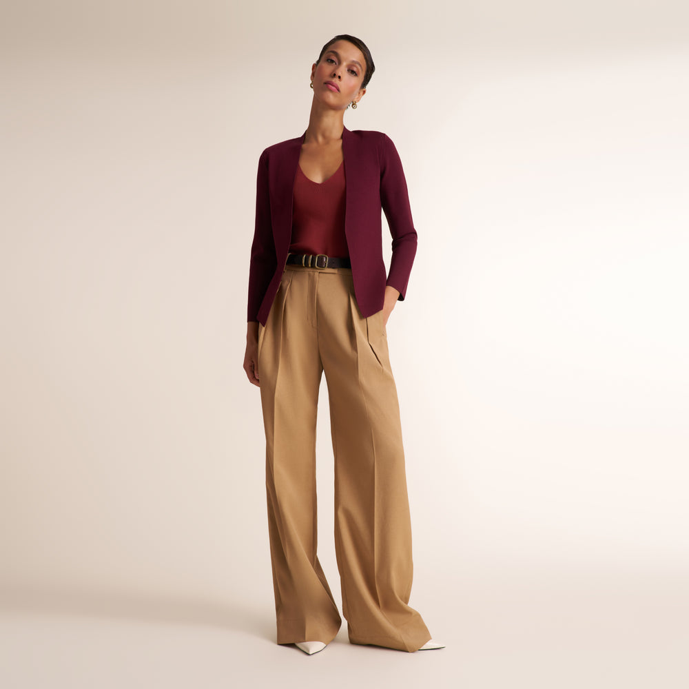 A model stands against a plain beige studio background wearing a dark burgundy long-sleeve knit top, a matching dark burgundy cardigan, high-waisted wide-leg tan trousers, and white pointed shoes.