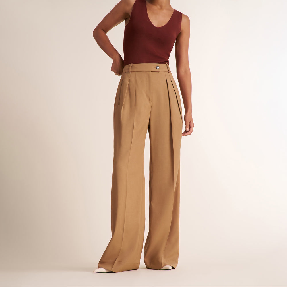 A model wearing high-waisted, wide-leg tan trousers with pleats and a maroon sleeveless top, standing against a plain light beige studio background.