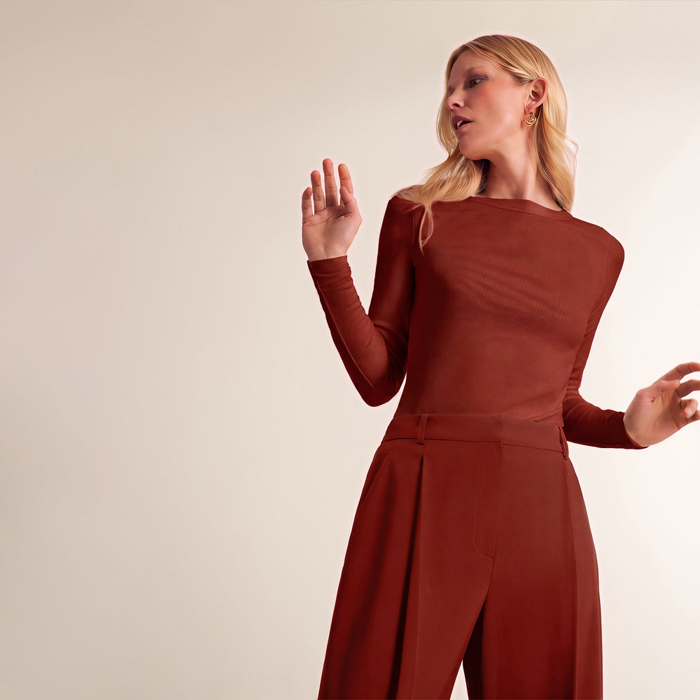 A woman modeling a rust-brown long-sleeve jumpsuit against a plain beige studio background.