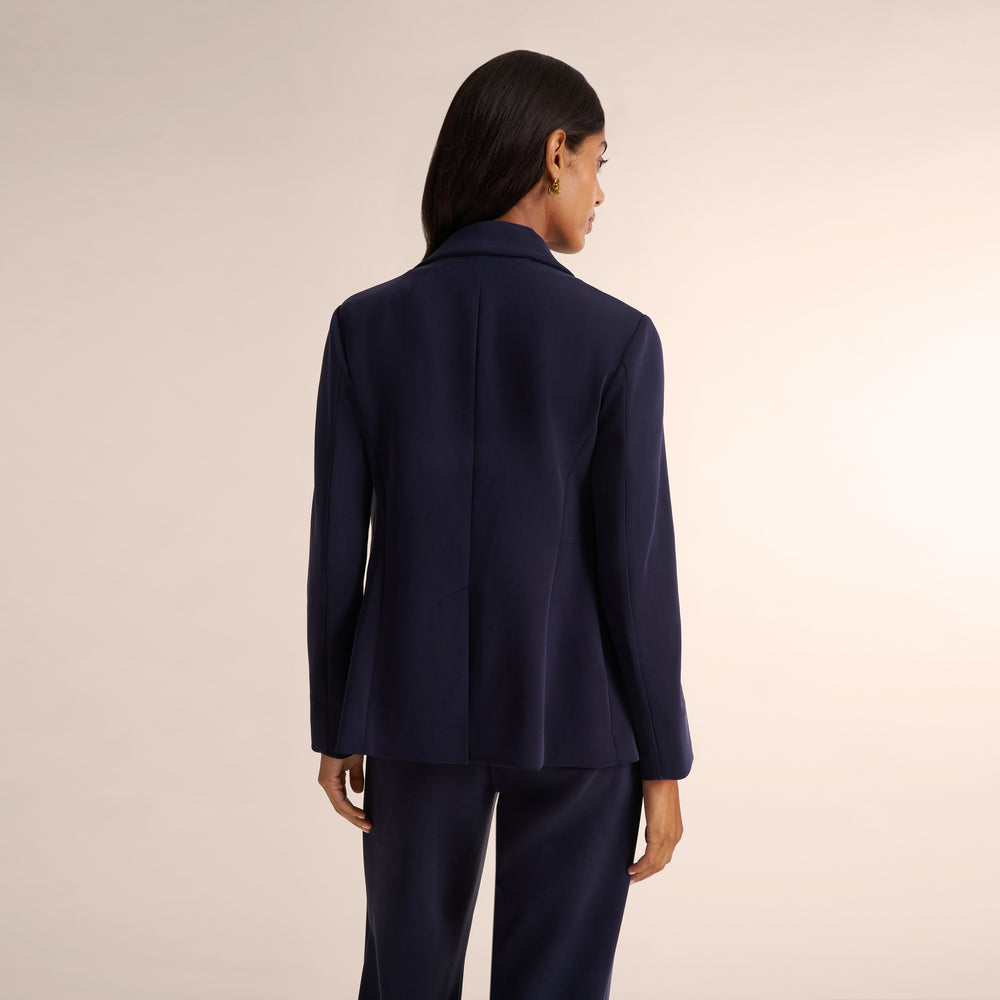 A model shown from the back wearing a dark navy blue tailored blazer and matching trousers against a plain light beige studio background.