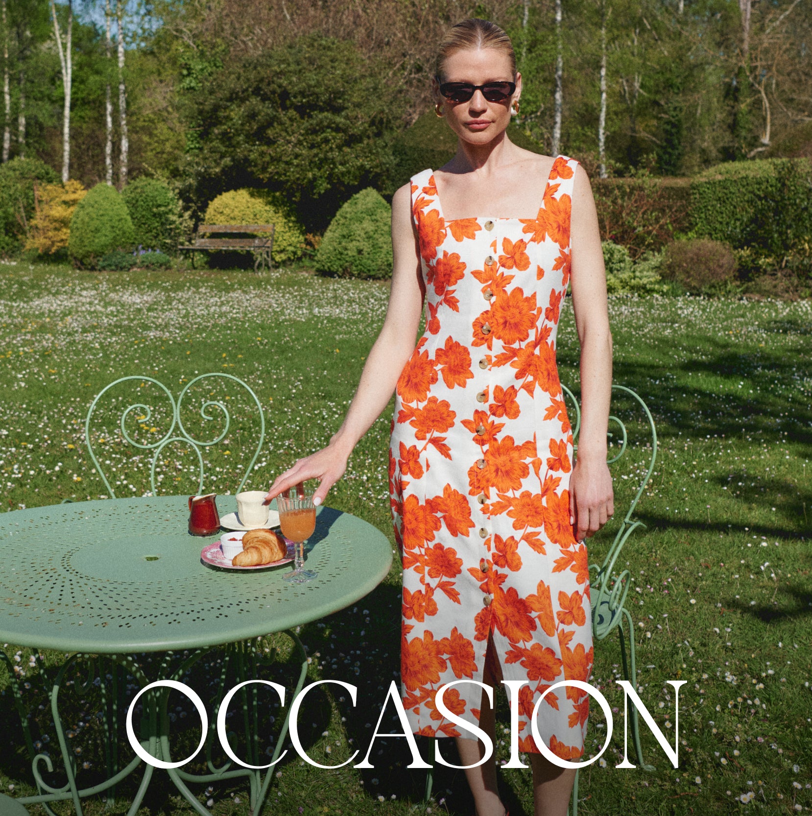 A woman wearing a sleeveless white dress with a bold orange floral pattern stands in a sunny garden next to a light green metal table. The brand name 'OCCASION' is displayed in white serif text at the bottom of the image.