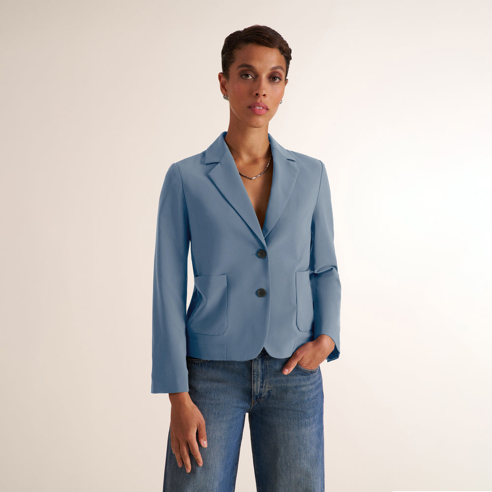 A model wearing a light blue tailored blazer with notched lapels and two front buttons, paired with blue jeans, against a plain light beige studio background.