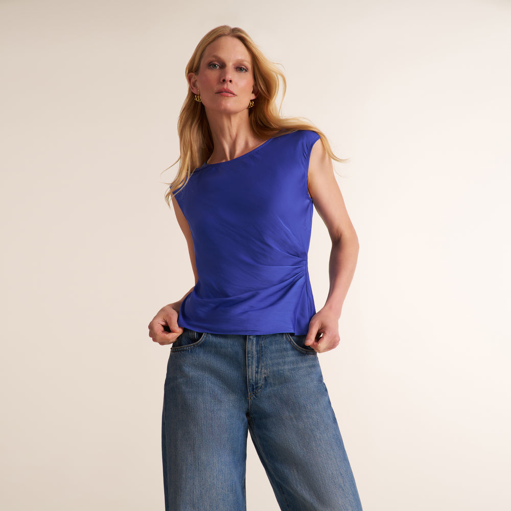 A woman modeling a sleeveless, one-shoulder royal blue top with gathered detailing, paired with blue jeans, against a plain light beige studio background.