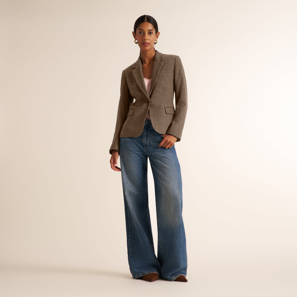 A model stands against a warm beige studio background wearing a brown textured blazer, wide-leg blue jeans, and brown shoes.