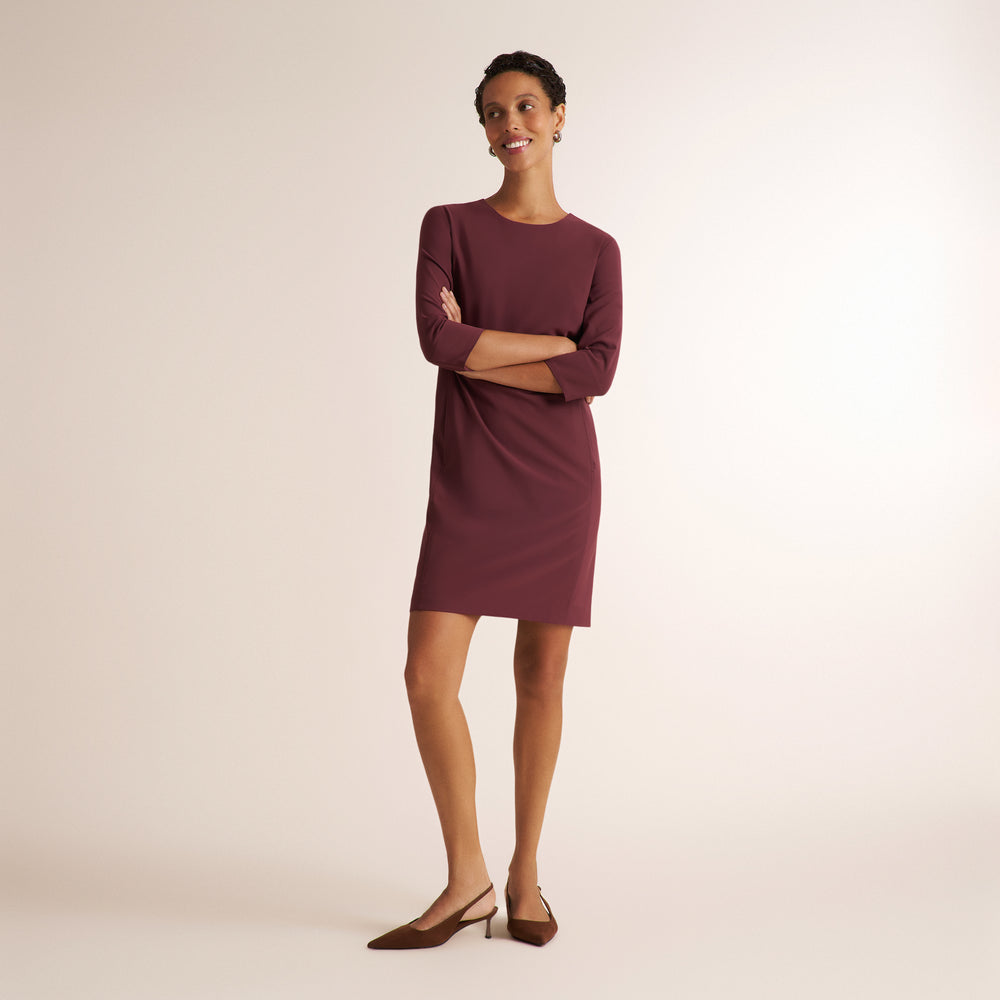 A woman modeling a long-sleeve, knee-length burgundy dress with a crew neckline, paired with brown slingback heels, against a plain beige studio background.