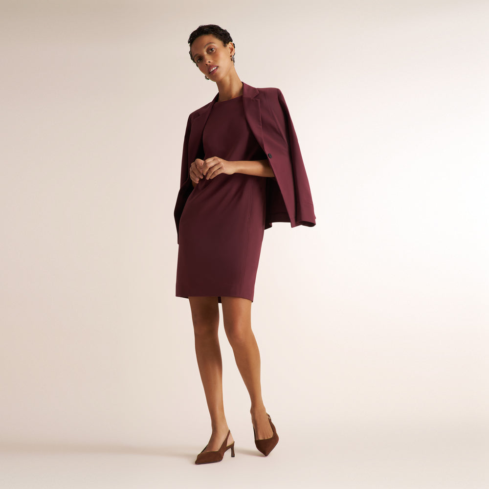 A woman models a short, bodycon dress in deep burgundy with a matching draped cape-style jacket, paired with brown pointed-toe heels, against a plain light beige studio background.