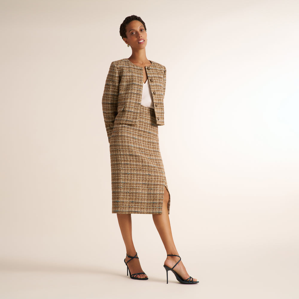 A model stands in a studio wearing a matching tweed jacket and midi skirt set in a brown and beige plaid pattern, styled with a white top and black strappy heels.