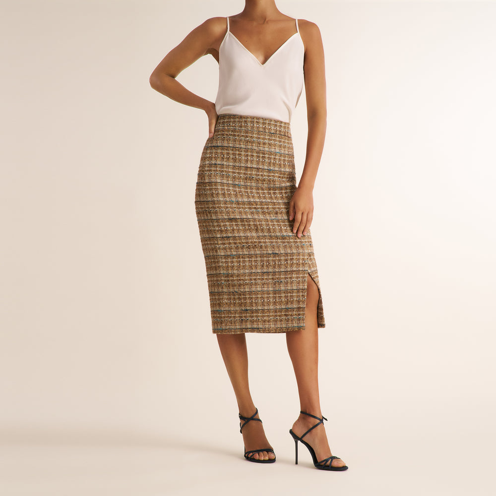 A model wearing a brown and beige tweed pencil skirt with a side slit, paired with a white camisole top and black strappy high-heeled sandals, against a solid light beige background.