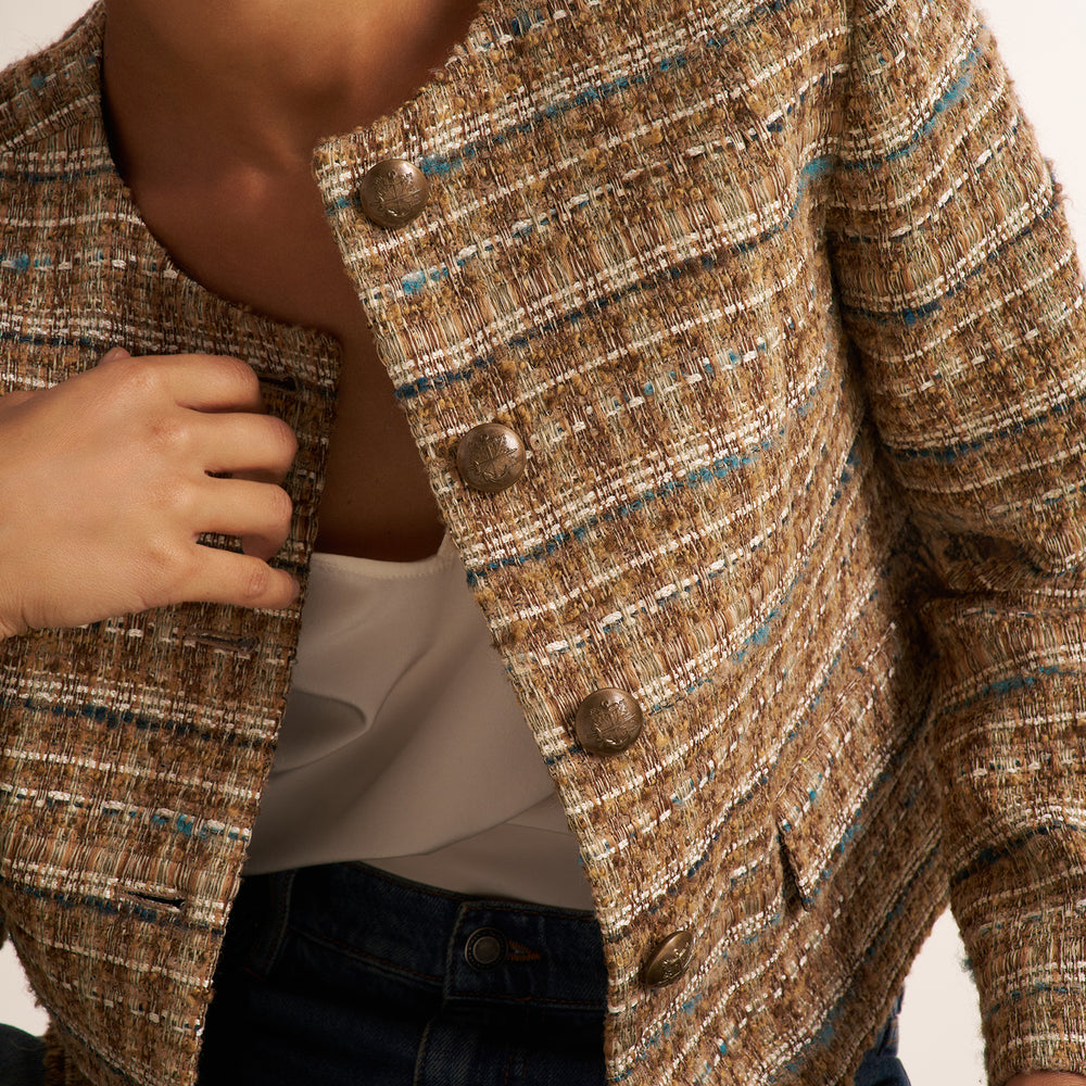 Close-up of a person wearing a textured tweed jacket in a warm brown, beige, and blue-gray mix, styled over a white top and dark blue jeans.