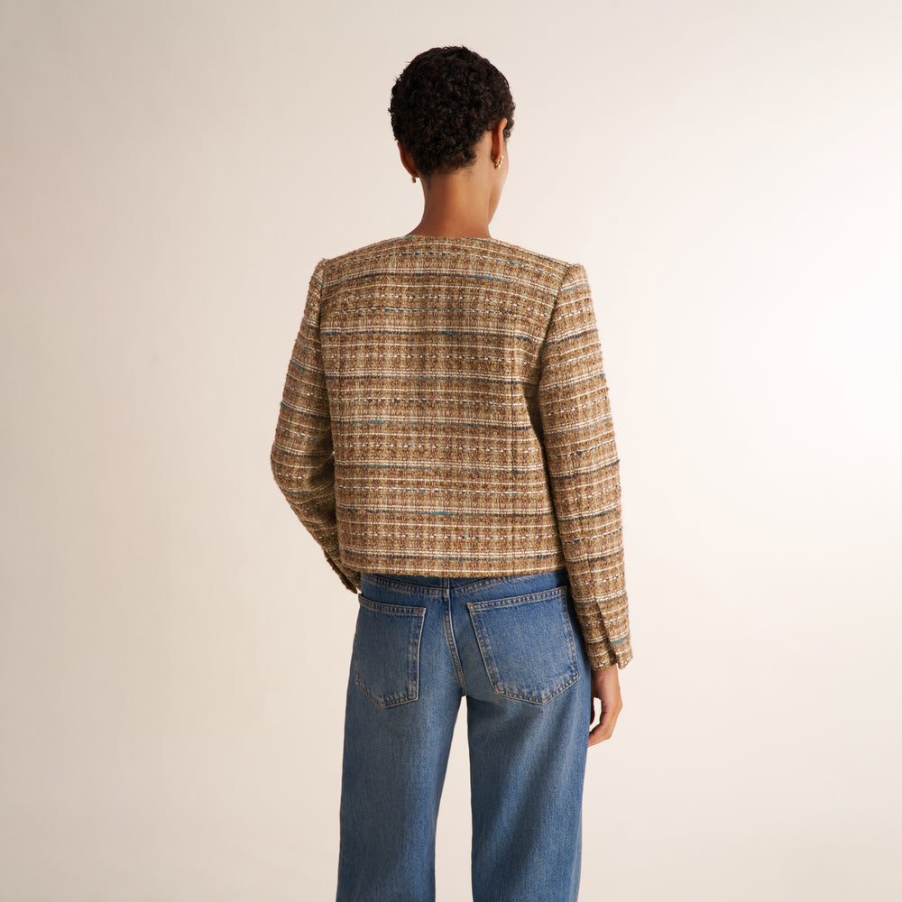 A model shown from the back wearing a cropped tweed jacket in a brown and beige pattern, paired with blue jeans, against a plain beige background.