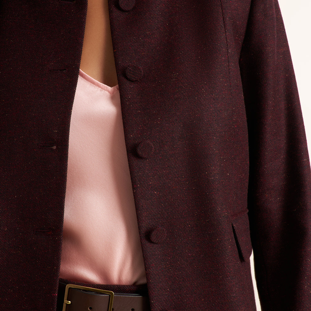 Close-up of a dark burgundy tweed blazer with dark buttons, worn over a light pink satin top and a brown belt with a gold buckle.
