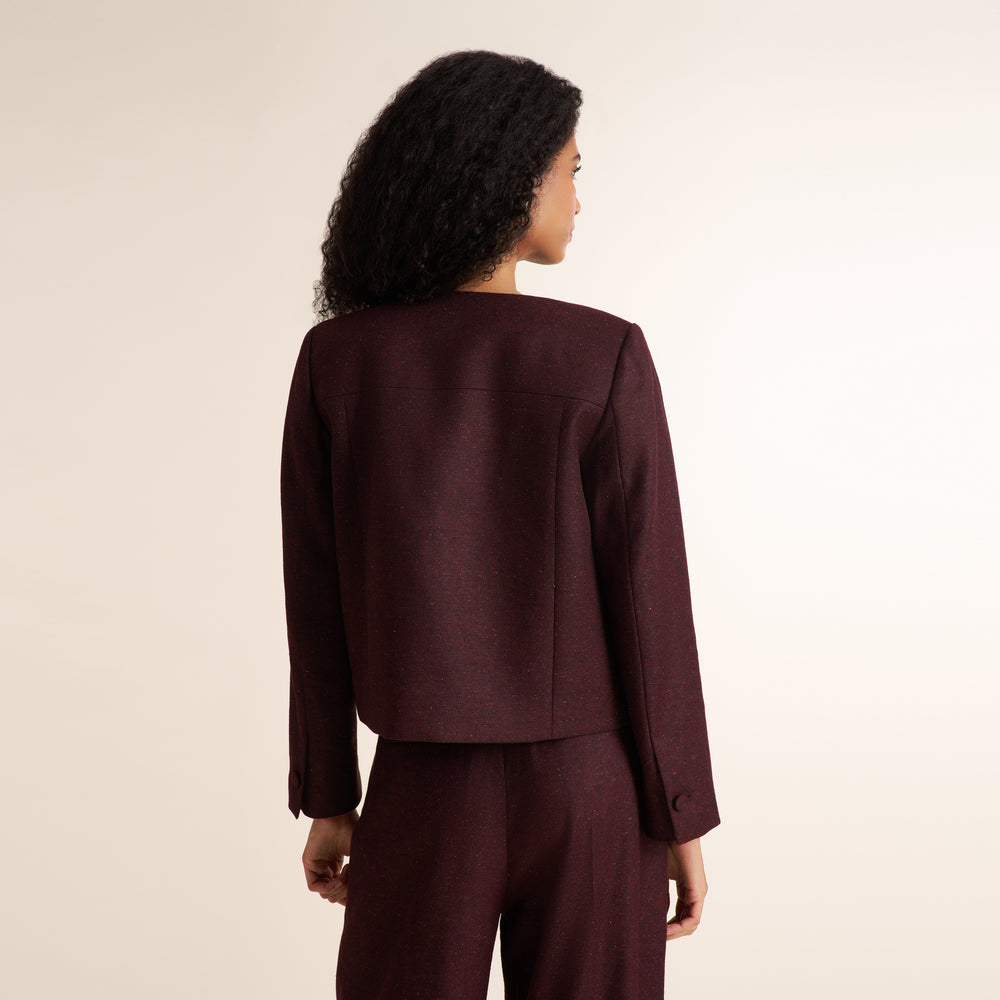 A woman shown from the back wearing a dark burgundy cropped blazer and matching trousers against a plain light beige studio background.