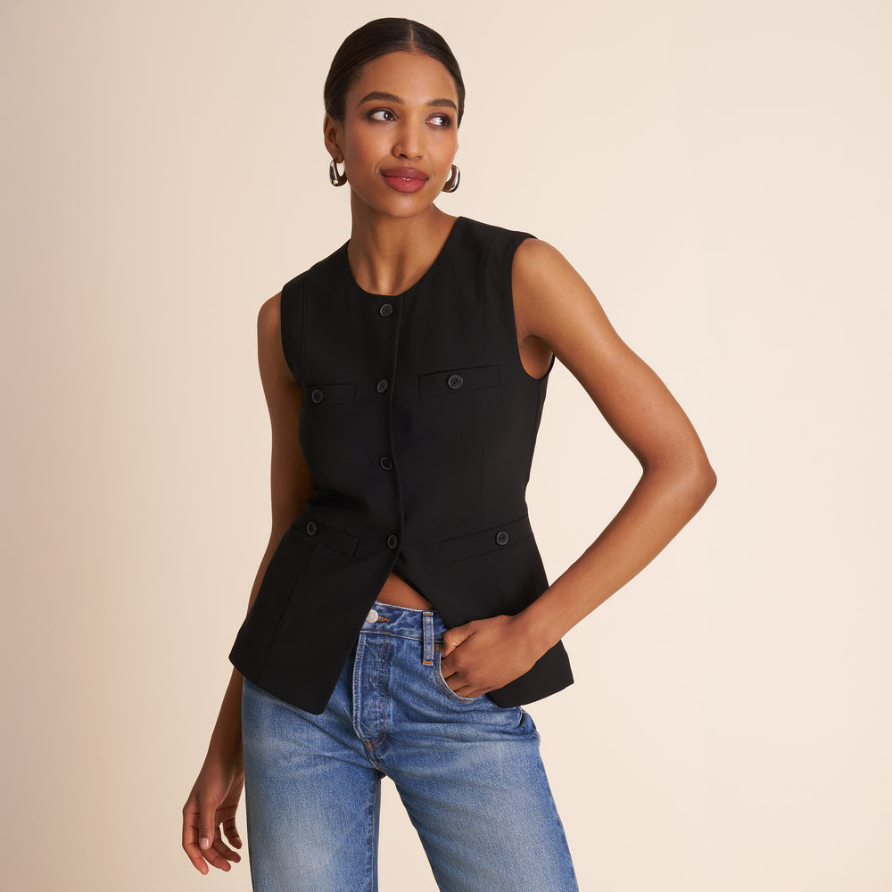 Woman wearing a black sleeveless blazer and blue jeans against a beige background