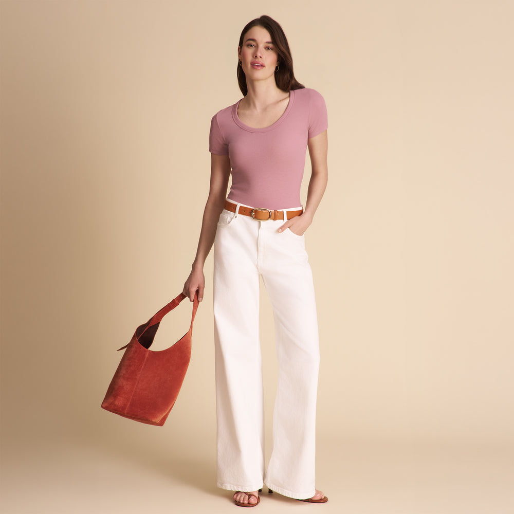 Woman wearing a pink top and white pants, holding an orange bag against a beige background