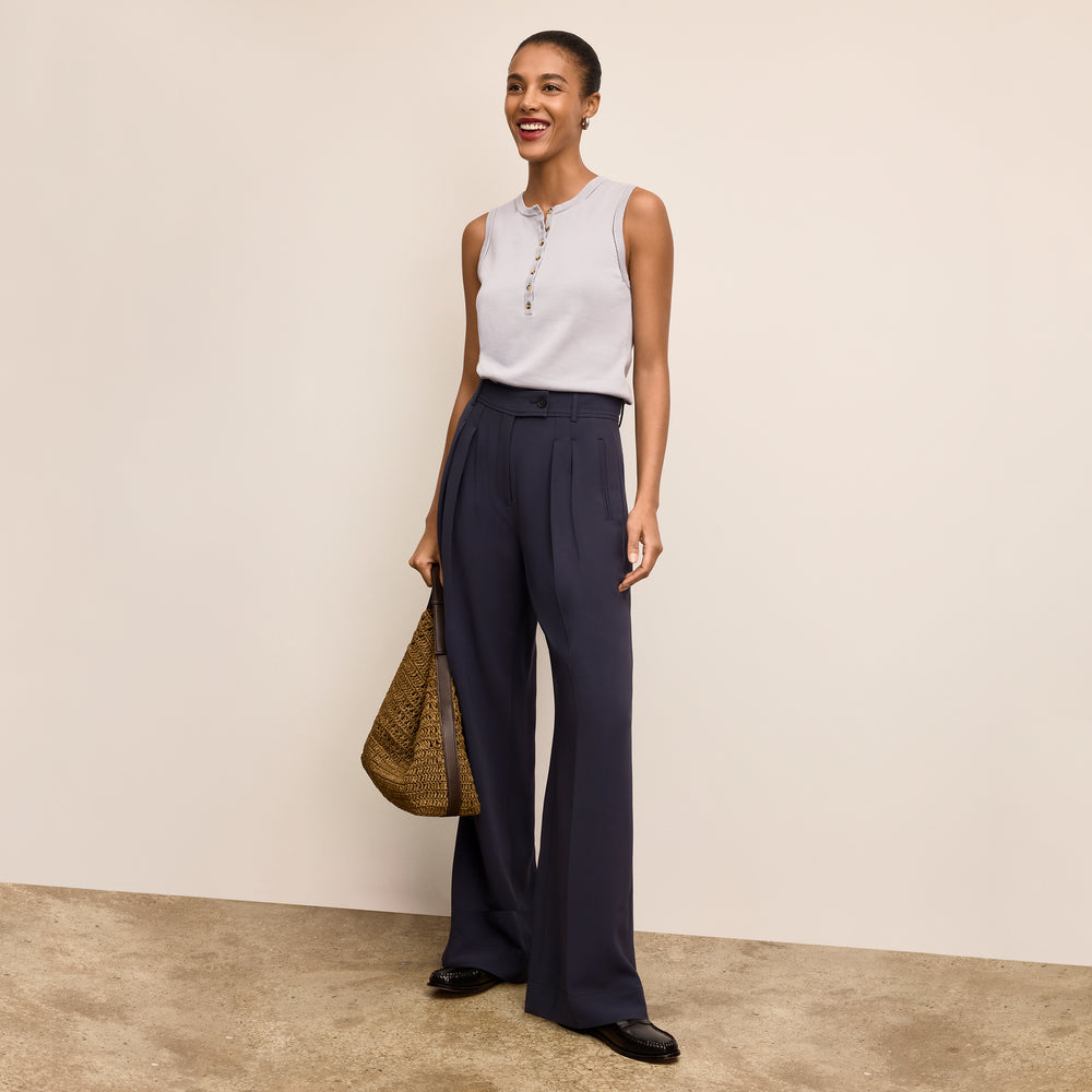 Woman wearing a sleeveless top and wide-leg pants, holding a bag against a plain background