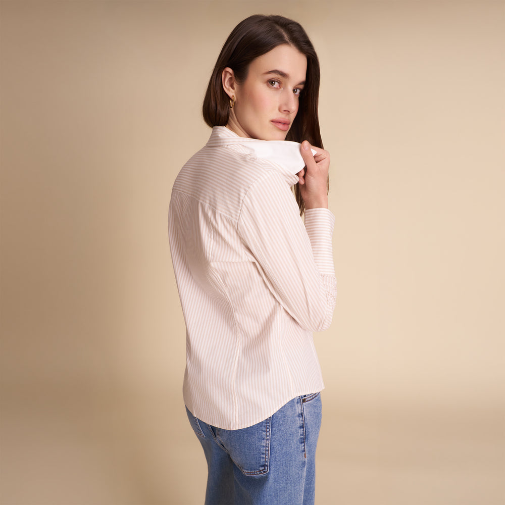 A woman wearing a light pink and white vertically striped long-sleeve blouse with a high folded collar, paired with blue jeans, photographed against a solid beige studio background.