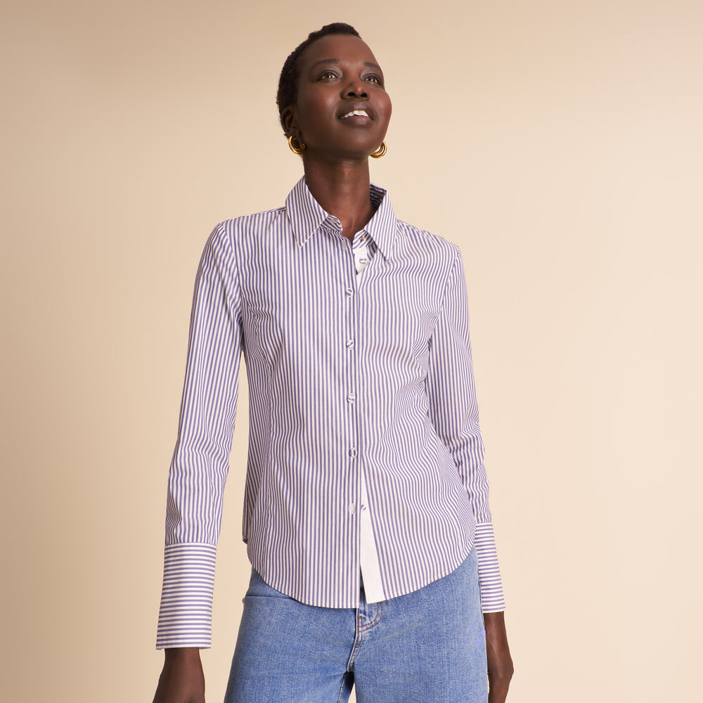 A woman wearing a long-sleeve button-up shirt with thin vertical navy and white stripes, paired with light blue jeans, standing against a solid beige background.