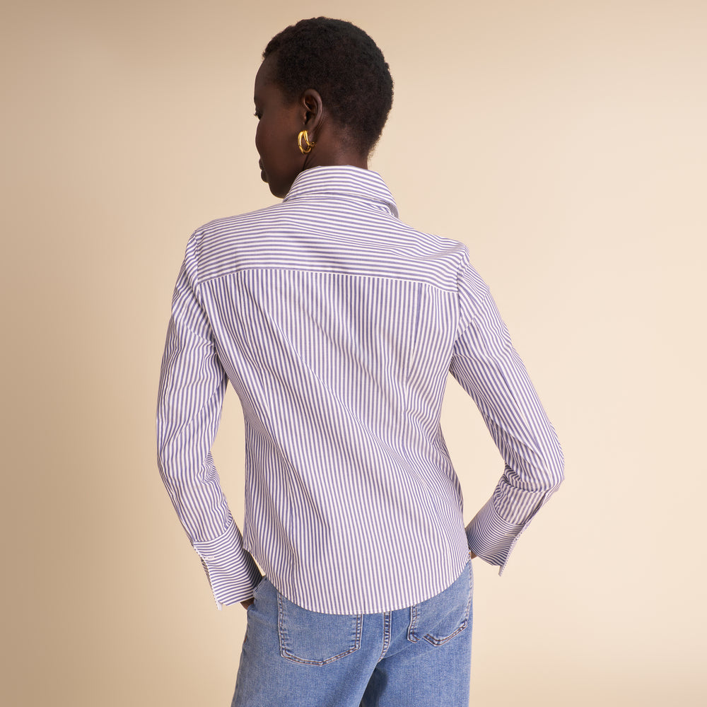 A woman shown from the back wearing a light blue and white vertically striped button-down shirt with light blue jeans, set against a solid beige studio background.