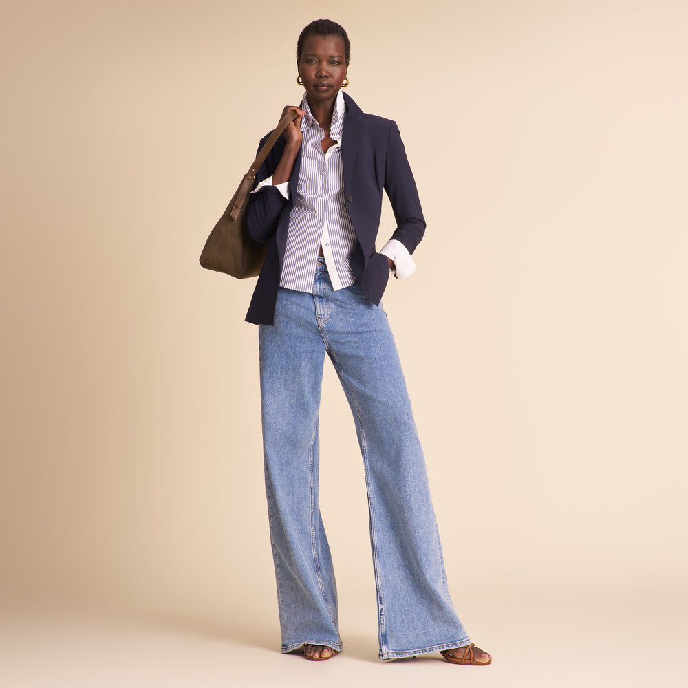 Woman wearing a navy blazer, white shirt, and blue jeans holding a brown handbag on a beige background