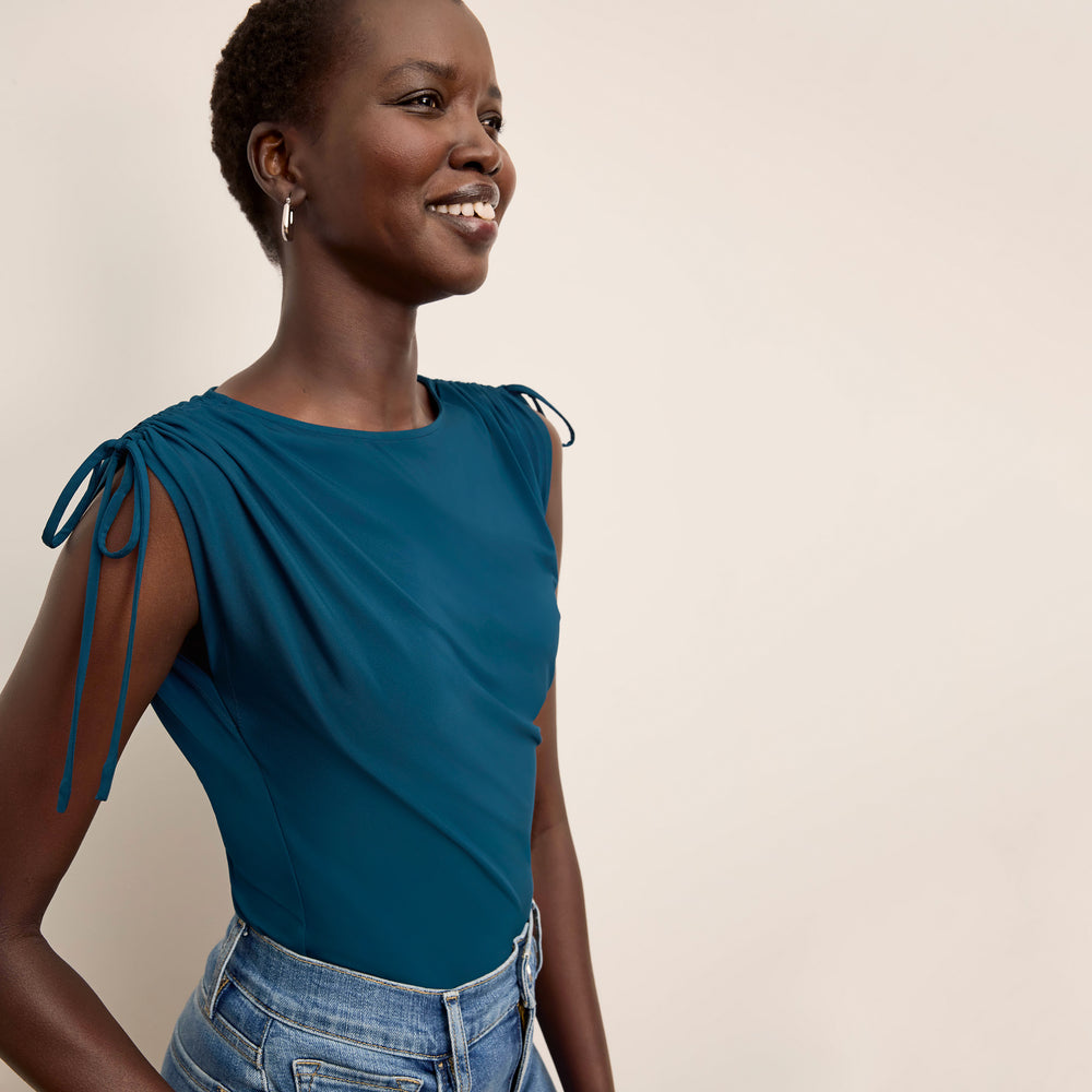 Woman wearing a teal top with tied sleeves against a beige background