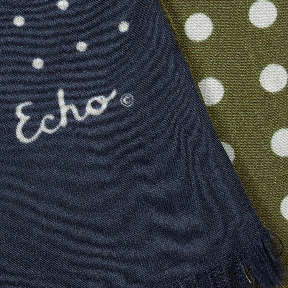 Close-up of a woven scarf with fringe, showing a navy section with white embroidered 'Echo' script and white dots, next to an olive green section with large white polka dots.