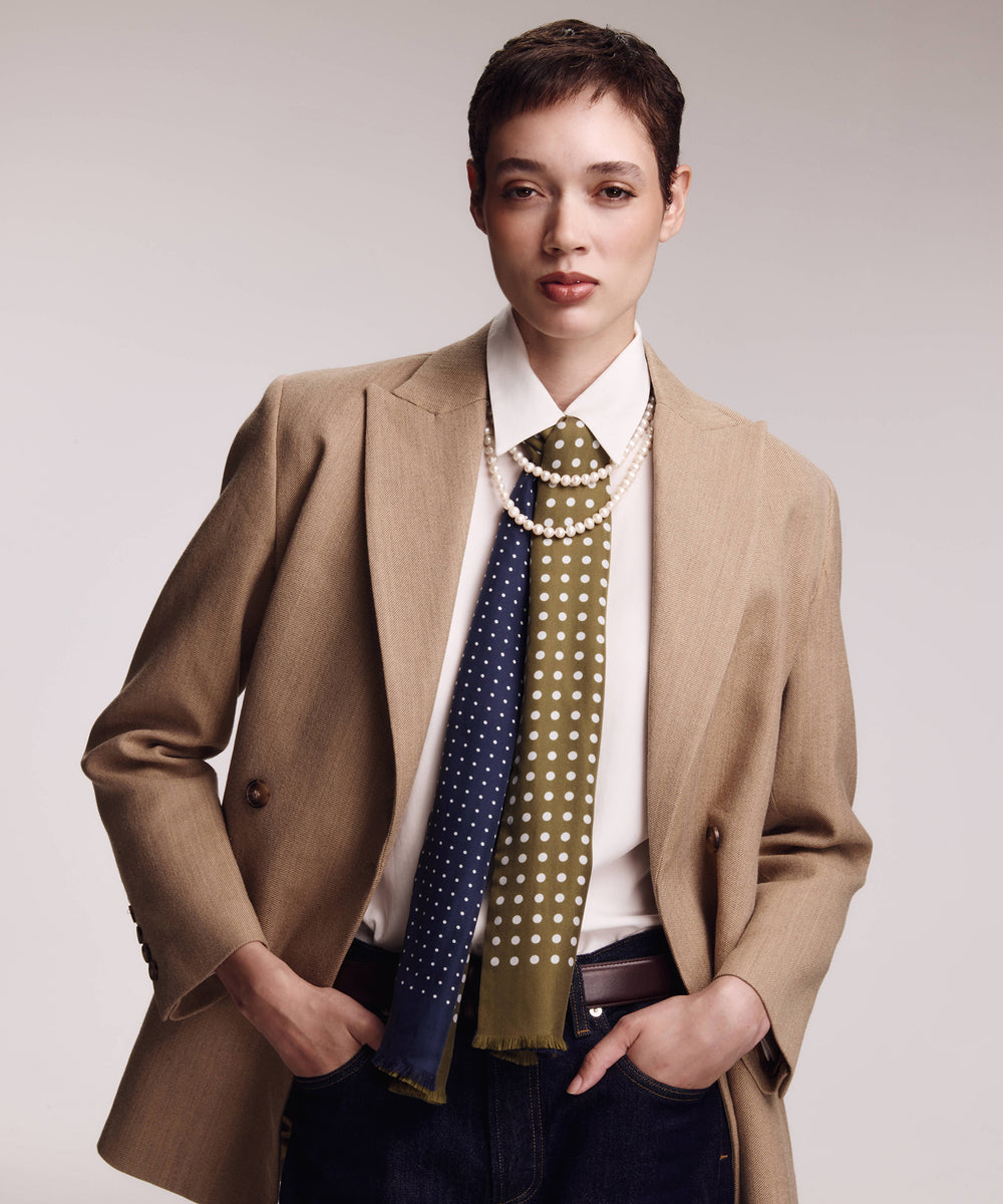 A model wearing a beige blazer over a white shirt, styled with two patterned neckties (one navy blue with white dots, one olive green with white dots) and layered with a pearl necklace.