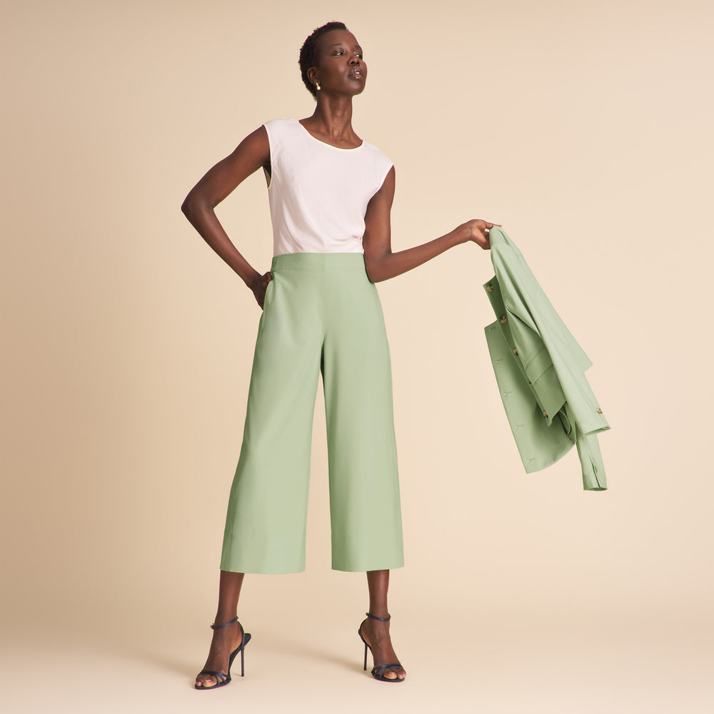 Woman wearing a white top and light green pants holding a matching jacket against a beige background