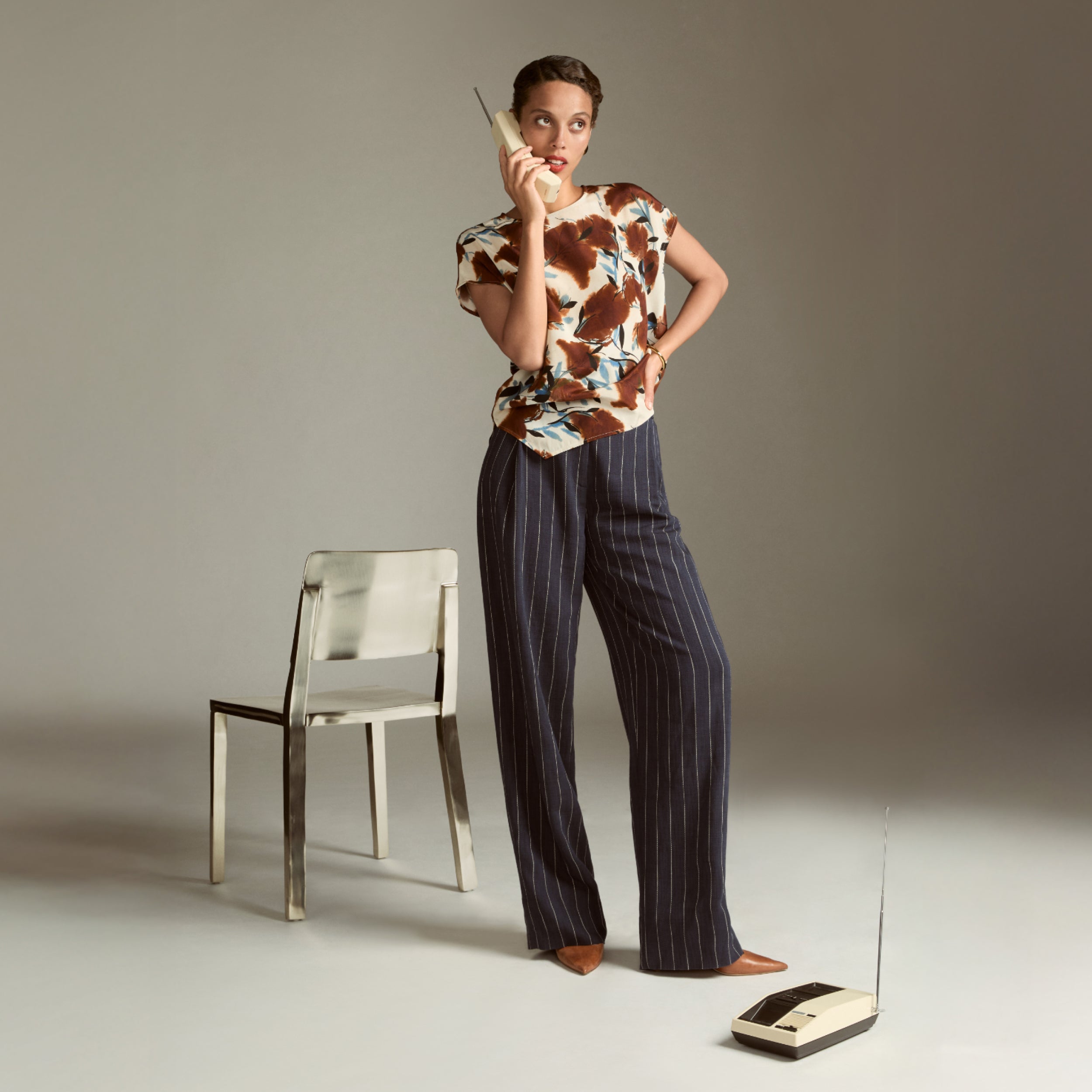 Woman in a patterned top and striped pants standing next to a chair and old-fashioned phone on a plain background | Exclude