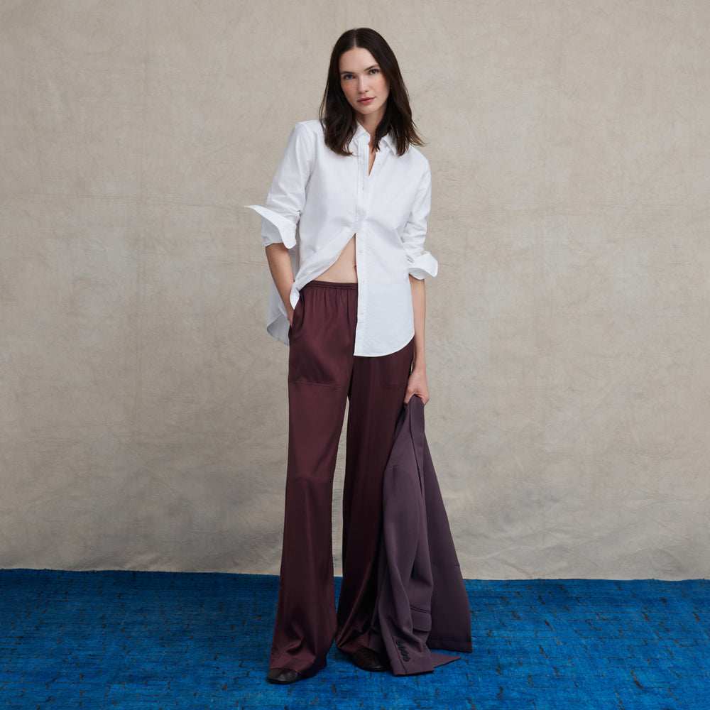 A woman stands against a textured beige backdrop wearing a white button-up shirt and wide-leg burgundy trousers, with a dark purple draped fabric held in one hand.